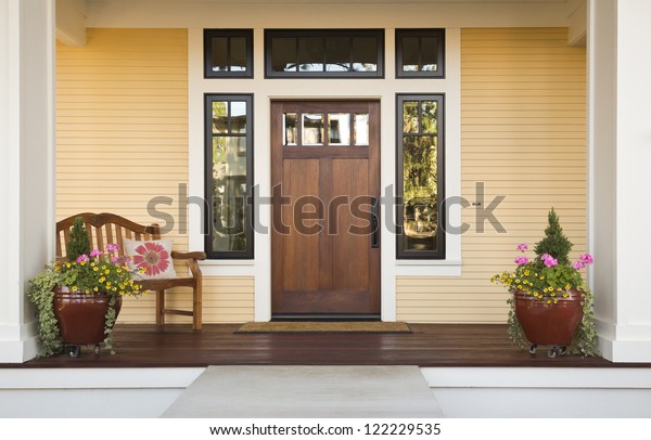 Wooden front door of\
a home. Front view of a wooden front door on a yellow house with\
reflections in the window and a wide view of the porch and front\
walkway. Horizontal\
shot.