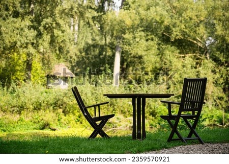 Wooden garden furniture,  a table and two chairs, are constrasting against the green grass and trees of an hidyllic garden, with lots of free (negative) space.