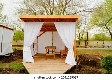 Wooden Gazebo With White Curtains For Lunch In Nature. Construction And Garden Furniture For A Country House