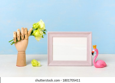 Wooden hand of manikin hold tender flowers, figurine of pink flamingo, photo frame on blue background and white wooden table Mock up Home decor, interior concept Scandinavian style Part of room design - Powered by Shutterstock