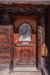 A Wooden House With An Old Wooden Door. In Front Of The Window Is A Metal Frame In The Shape Of A Bird. An Old-fashioned Broom Hangs Next To The Door. Switzerland.