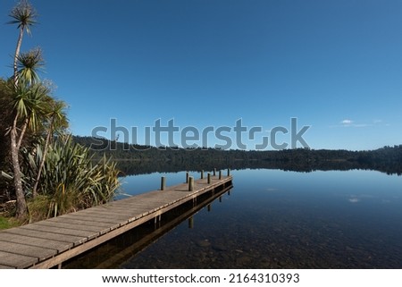 A wooden jetty reflected in a flat calm Lake Ianthe under a clear, blue sky.. West Coast, New Zealand.