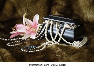 A Wooden Jewelry Box Overflowing With Pearl Jewelry, Photographed On A Faux Fur Underlay.  Pink Lilies Are Placed To The Side Of The Box As An Accent.