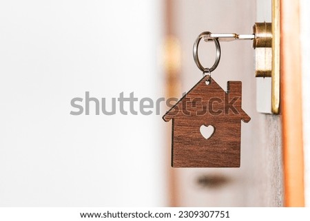 Wooden key chain in the shape of a house with a heart-shaped hole in the center hanging from a key inserted in the lock of a front door of a house.