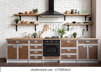 Wooden kitchen facade in apartment with modern interior, new furniture, gas stove, built in oven equipment, cooking hood, kitchenware supplies near sink and houseplant in flower pot