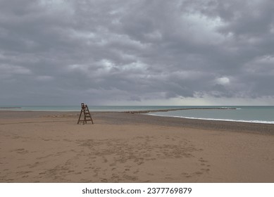 Wooden lifeguard chair on lonely beach. Cloudy sky, sandy beach, sky with storm clouds, turquoise sea, pebble jetty - Powered by Shutterstock