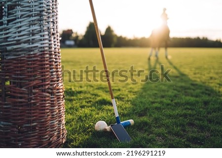 Wooden mallet ball and mobile phone left on grass field by polo player during training along with wicker basket at sunset with rider on background