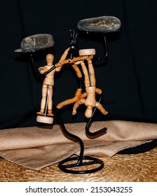 Wooden manikins on under a rock posing upside down and right side up in different positions. The manikin is handing on a black figurine balancing the manikins.