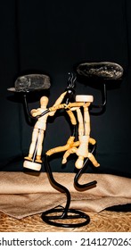Wooden manikins are under a rock posing upside down  in different positions . The manikins are handing on a black figurine balancing itself.
				
