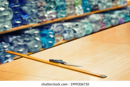 Wooden Metre Ruler And Tailor Scissors Lie On The Table In Fabric Shop. Different Fabric And Textile On Background. Tools For Measuring And Cutting Textile.