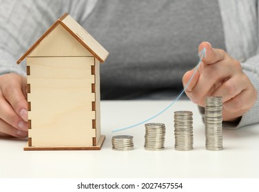 Wooden Model Of A House, A Stack Of Coins And An Ascending Graph. The Man Sits At The Table. Higher Property Prices, Higher Rents And Higher Mortgage Interest Rates