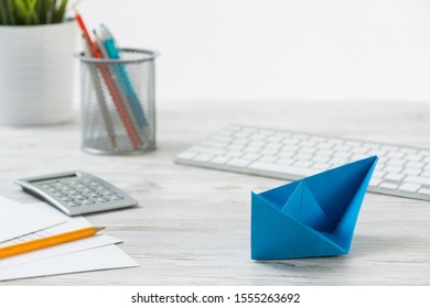 Wooden Office Desk With Deep Blue Origami Boat. Individual Banking And Money Saving. Flat Lay Office Workplace With Plant In Pot, Computer Keyboard And Calculator. Creative Company Marketing Concept