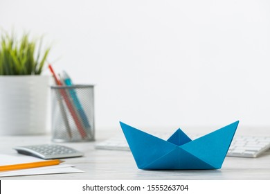 Wooden Office Desk With Deep Blue Origami Boat. Individual Banking And Money Saving. Flat Lay Office Workplace With Plant In Pot, Computer Keyboard And Calculator. Creative Company Marketing Concept