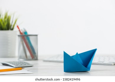 Wooden Office Desk With Deep Blue Origami Boat. Individual Banking And Money Saving. Flat Lay Office Workplace With Plant In Pot, Computer Keyboard And Calculator. Creative Company Marketing Concept