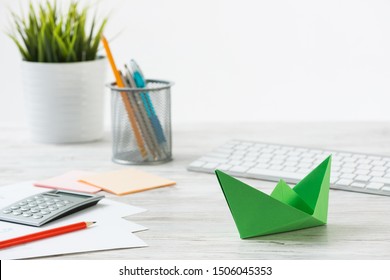 Wooden Office Desk With Green Origami Boat. Individual Investment And Money Saving. Flat Lay Office Workplace With Plant In Pot, Computer Keyboard And Calculator. Green Technologies And Innovations