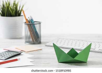 Wooden Office Desk With Green Origami Boat. Individual Investment And Money Saving. Flat Lay Office Workplace With Plant In Pot, Computer Keyboard And Calculator. Green Technologies And Innovations