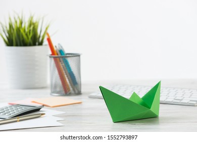 Wooden Office Desk With Green Origami Boat. Individual Investment And Money Saving. Flat Lay Office Workplace With Plant In Pot, Computer Keyboard And Calculator. Green Technologies And Innovations