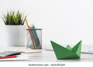 Wooden Office Desk With Green Origami Boat. Individual Investment And Money Saving. Flat Lay Office Workplace With Plant In Pot, Computer Keyboard And Calculator. Green Technologies And Innovations