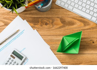 Wooden Office Desk With Green Origami Boat. Individual Investment And Money Saving. Flat Lay Office Workplace With Plant In Pot, Computer Keyboard And Calculator. Green Technologies And Innovations