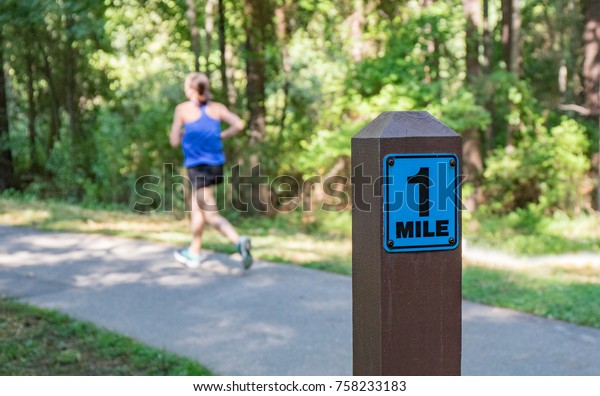 A wooden one mile
marker post in the foreground with a woman running in the
background on a cement pathway.  Shallow depth of field so the
background is out of focus.  