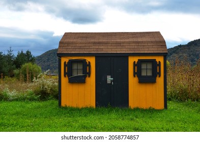 Wooden Outdoor Storage Shed With Two Windows, Located In Backyard Of Countryside. Yellow Color With Black Window Frames. American Traditional Style Shed.