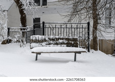 A wooden park bench on a street corner with a wrought iron fence and a white wooden building in the background. The seat is covered in fresh white fluffy snow and ice. The ground is snow covered. 