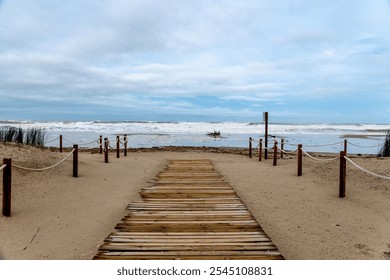 Wooden path leads from sandy beach to stormy sea, flanked by posts; driftwood and seaweed scattered on shore after storm. - Powered by Shutterstock