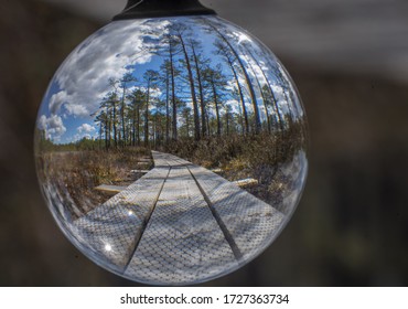 Wooden Path In Raised Bog Via Crystal Ball. Walking Road Planks Going Straight. Bokeh Background. Full World In Small Glass Sphere.  Early Spring Weather, Blue Bright Sky. Estonia, Baltic, Europe.