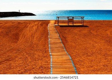 Wooden pathway leading to ocean on a sandy beach. The pathway is bordered by blue lines and extends from the foreground to the middle. In the background, the calm blue sea with rocky outcrop - Powered by Shutterstock