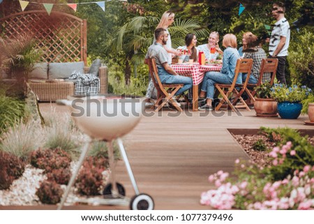 Wooden patio in the garden with a grill standing in the front and happy young people gathered around a table full of food during summer brake meeting