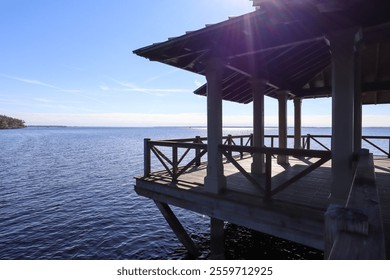 A wooden pavilion extends over the calm water, with sunlight casting bright reflections on the surface, offering a peaceful and scenic view of the tranquil lakeside under a clear blue sky. - Powered by Shutterstock