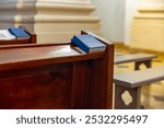 Wooden pew in a church. There is a hymnal book on the pew. The pew is next to a second pew in the background. Both pews are made of wood.