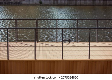 A wooden pier by a river, pond, lake with dark calm water. A place near a shore for mooring boat, boats, yachts. Location for walks of city dwellers, relaxation outdoors. Black metal fence, fencing