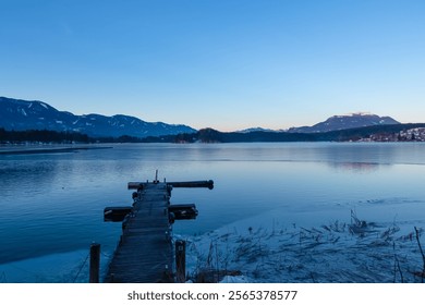 Wooden pier extends into partially frozen Lake Faak at sunset in Carinthia, Austria. Scenic view of high snow capped mountains peaks of Karawanks, Julian Alps and Dobratsch. Winter in Austrian Alps - Powered by Shutterstock