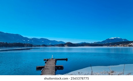 Wooden pier extends into partially frozen Lake Faak in Carinthia, Austria. Panoramic view of surrounding snow capped mountains peaks of Karawanks, Julian Alps and Dobratsch. Winter in Austrian Alps - Powered by Shutterstock