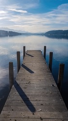 A Wooden Pier Going Into The Woerthersee In Poertschach In Carinthia, Austria. The Calm Surface Of The Lake Is Reflecting The Mountains, Sunbeams And Clouds. Clear And Sunny Day. Alps, Lake Woerth