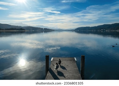 A wooden pier going into the Woerthersee in Poertschach in Carinthia, Austria. Flock of ducks on the pier. Calm surface of the lake is reflecting the mountains. Clear and sunny day. Alps, Lake Woerth
