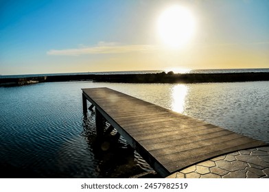 A wooden pier is on the water with the sun shining on it. The water is calm and the sky is blue, real image - Powered by Shutterstock