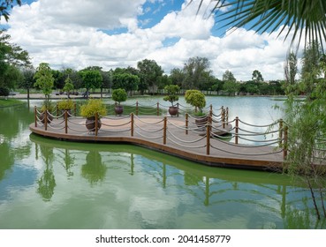 Wooden Pier With Plants At The Center Of A Lake Surrounded By Green Trees, Day With Blue Sky And Clouds, No People