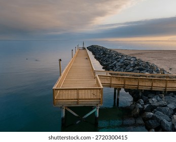 A wooden pier with a rectangular viewing platform extends into calm, glassy water along the North Shore of Long Island, New York. A rock jetty runs parallel to the pier, separating the water from a sandy shoreline. Soft, muted light illuminates layered clouds above Long Island Sound, creating gentle reflections on the still surface. The elevated perspective emphasizes strong leading lines, symmetry, and a quiet coastal atmosphere with no people present.