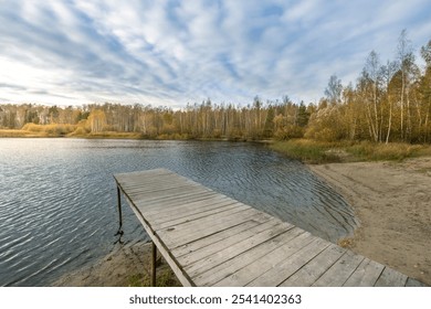 A wooden pier sits on the edge of a lake, with a cloudy sky overhead. The water is calm and still, and the trees in the background are bare and leafless. The scene is peaceful and serene - Powered by Shutterstock