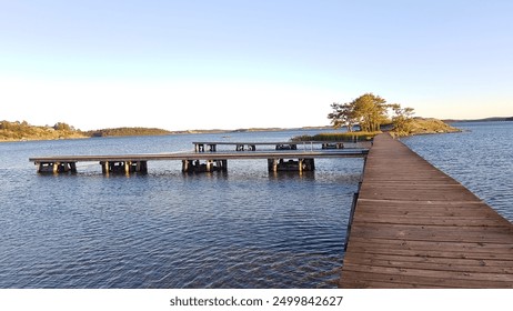 A wooden pier stretches across calm blue waters towards a small, tree-covered island. The scene captures the serene beauty of the landscape under a clear sky. - Powered by Shutterstock