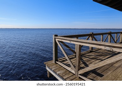 A wooden pier stretches over calm water, with a clear blue sky in the background. The wooden railing features a distinctive crisscross pattern, adding to the scenic waterfront view. - Powered by Shutterstock