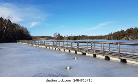 A wooden pier stretches over a frozen lake surrounded by trees and clear blue skies. Captures the serene and tranquil beauty of a winter landscape. - Powered by Shutterstock