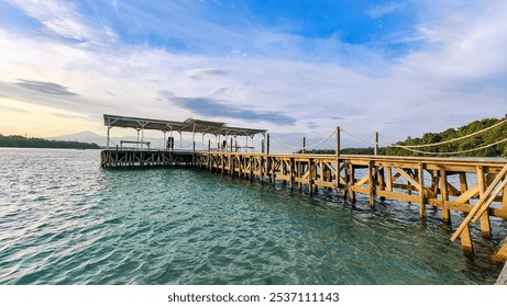 A wooden pier stretches over turquoise water, leading to a shaded structure with a view of the distant shoreline under a vibrant sky. - Powered by Shutterstock