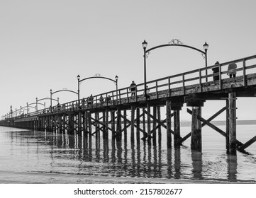 Wooden pier at White Rock, BC extends diagonally into image. Twinkling lights reflected in the sea. Black and white image: Beach and pier at sunset, Surrey, BC, Canada-February 22,2022