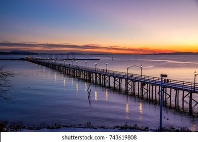 Wooden pier at White Rock, BC, Canada extends diagonally into image... its twinkling lights reflected in the sea.  The sun is setting with flaming reds and oranges over the Gulf and San Juan Islands

