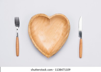 Wooden plate in shape of heart, table knife and fork on white background.