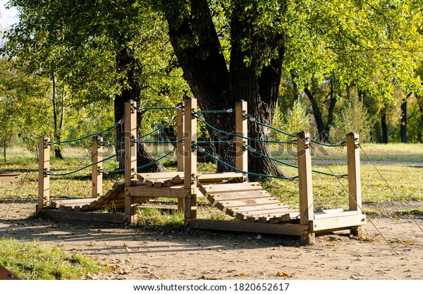 Wooden play bridge for children, located in the\
Park among the trees. Outdoor play facilities. Bridge made of\
wooden planks with rope\
railings.