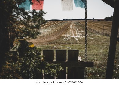 Wooden Porch Swing Looking Out At An Old Country Road Dusted With Snow In The Fall Beside An Evergreen Tree With Buddhist Prayer Flags Waving Overhead